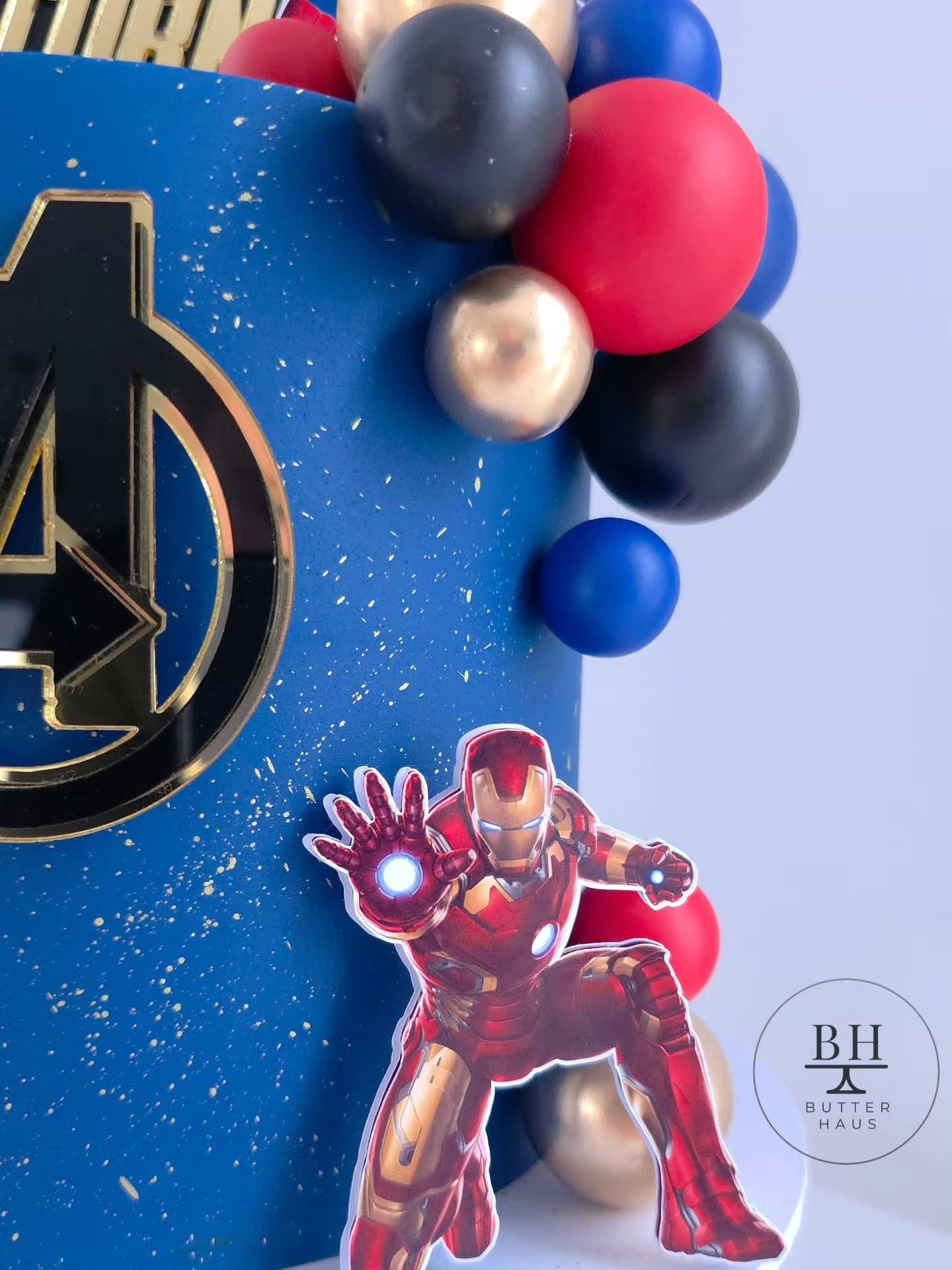 Close-up of gold mirror Avengers logo plaque and custom gold Name Turns Age topper on galaxy-effect buttercream cake