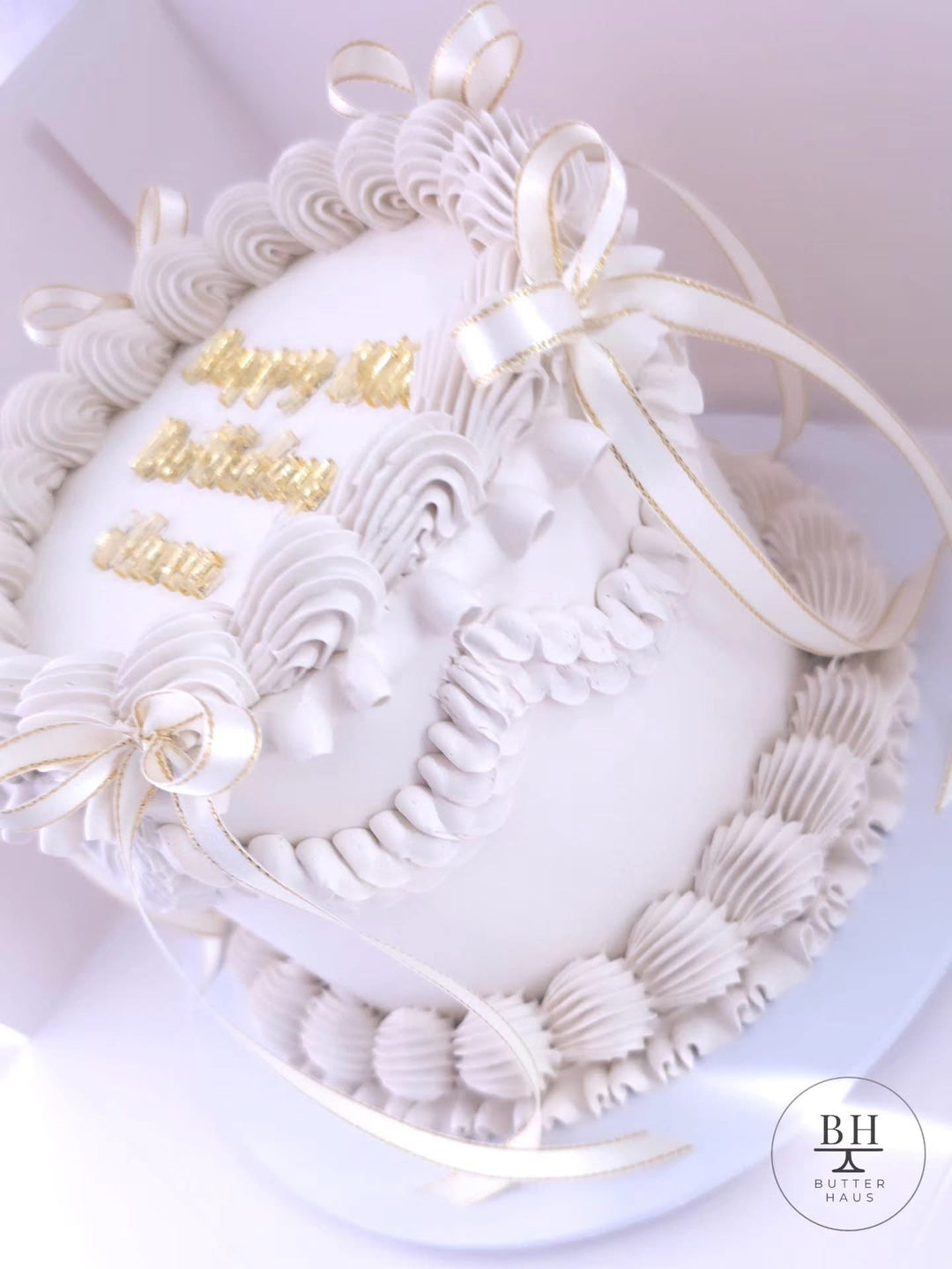 White vintage birthday cake with piped garlands, gold edged ivory bows and gold script topper
