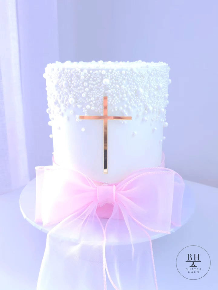 White cake covered in pearl sprinkles with a rose-gold cross and pale pink organza bow