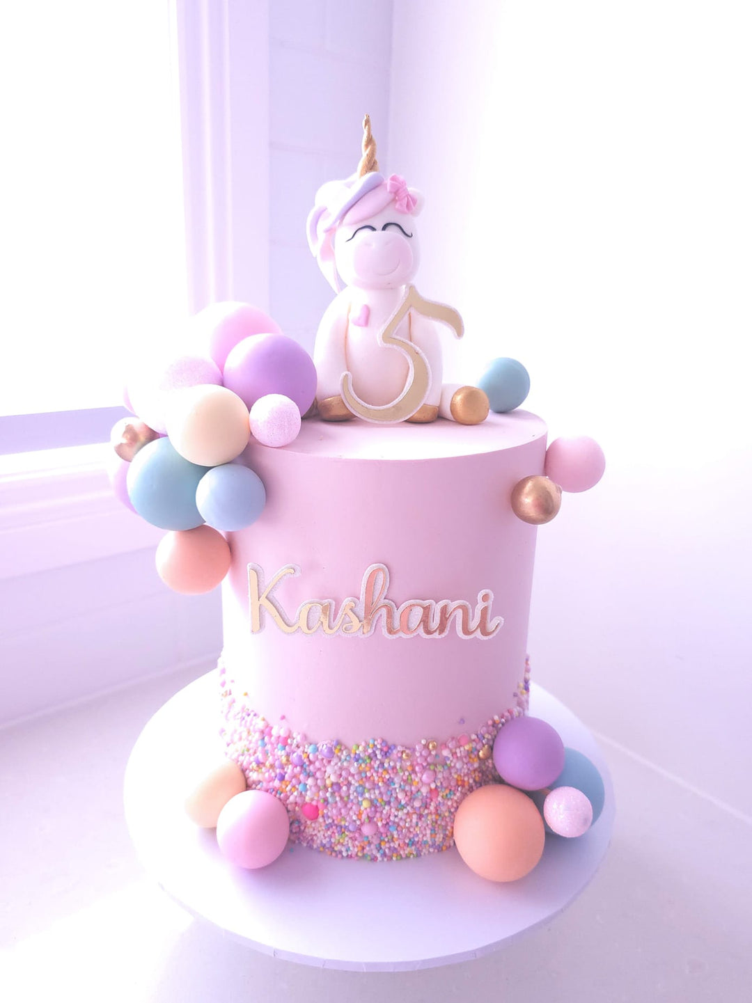 Unicorn Dreams birthday cake with pink buttercream, hand-modelled fondant unicorn topper and pastel chocolate spheres