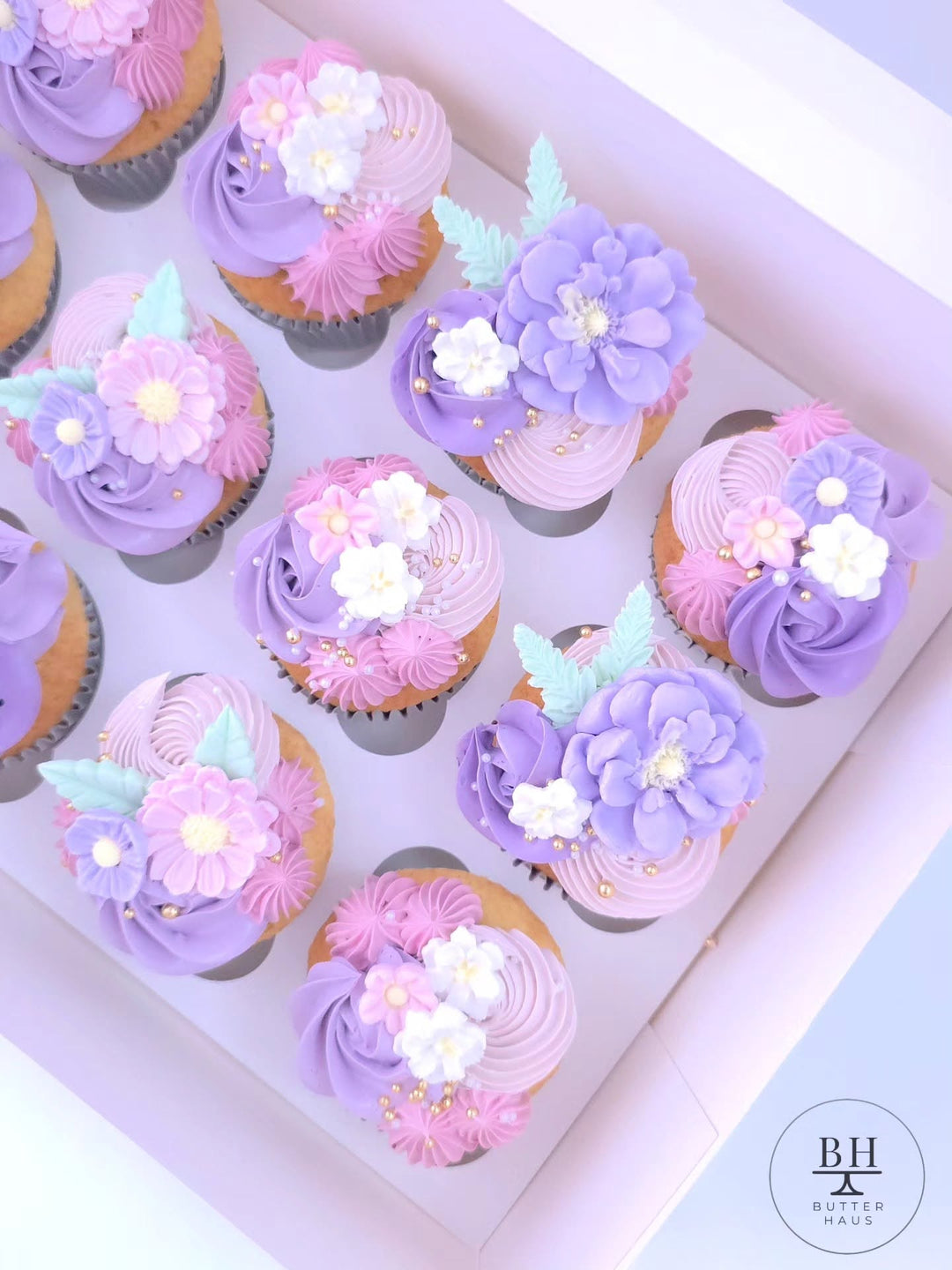 Box of purple and pink buttercream cupcakes topped with fondant flowers and pearls
